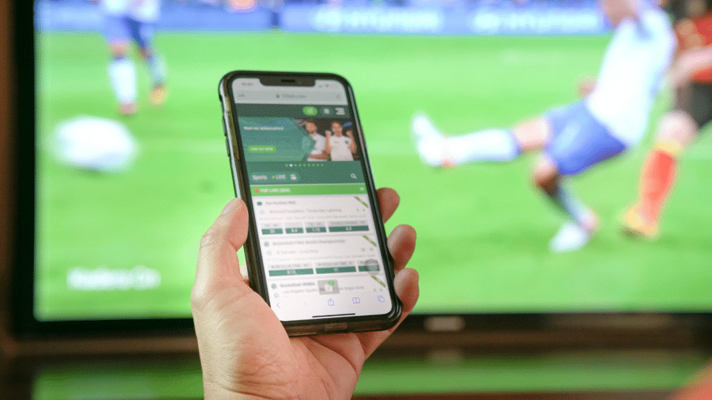 A person streaming a live sports match on a smartphone with a soccer game playing in the background on TV.