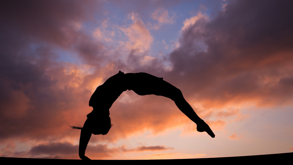 Silhouette of a gymnast performing a backbend under a vibrant sunset sky, symbolizing grace and strength in sports.